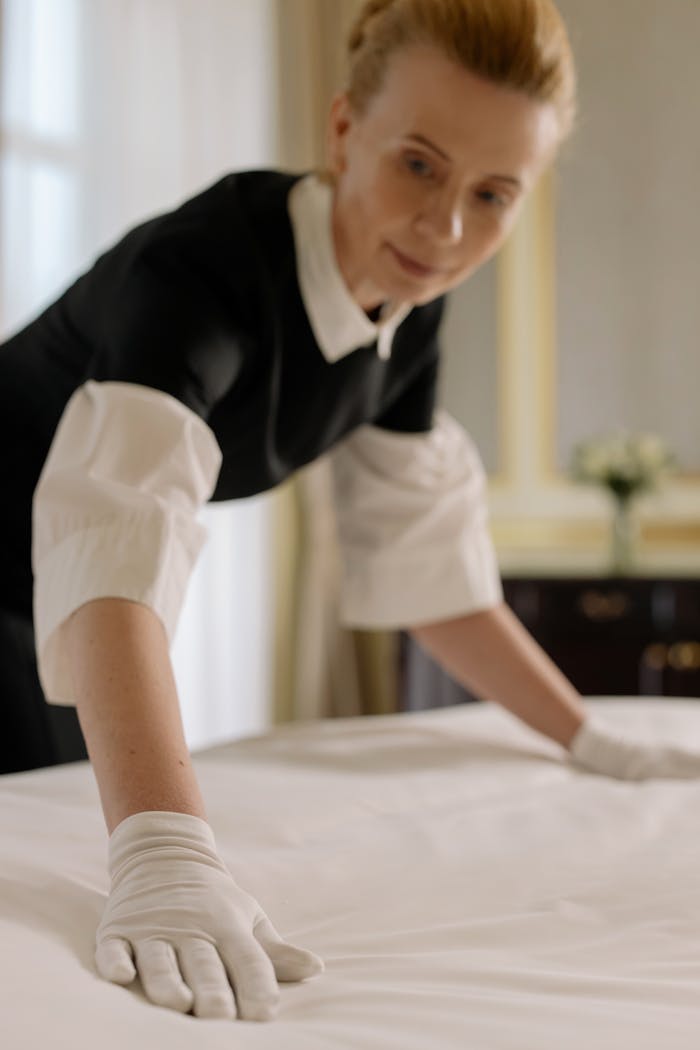 A housekeeper elegantly arranges bed sheets in a luxurious hotel room, showcasing hospitality expertise.